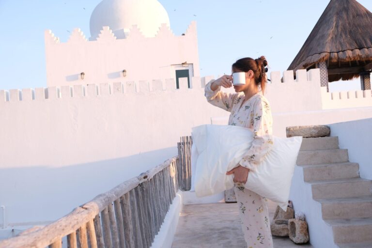 A woman in floral pajamas stands on a sunlit terrace, holding a large white pillow in one arm and sipping from a white mug with the other hand. Behind her are white stucco walls with decorative battlements, a domed roof, and a thatched gazebo-style roof. The scene is bright and serene, suggesting early morning.